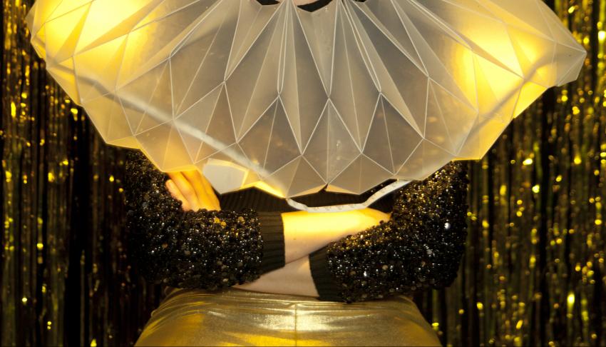 Milk Presents - A close-up image of a person wearing gold lamé leggings, a black sequened top and a white, opaque, plastic ruff around their neck and shoulders. They are sat in front of a gold glitter curtain. Their arms are crossed, their head and legs c
